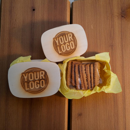 Zwei Holzkästchen mit gelasertem Logo, eines davon geöffnet und gefüllt mit einzeln verpackten Logokeksen auf rustikalem Holztisch