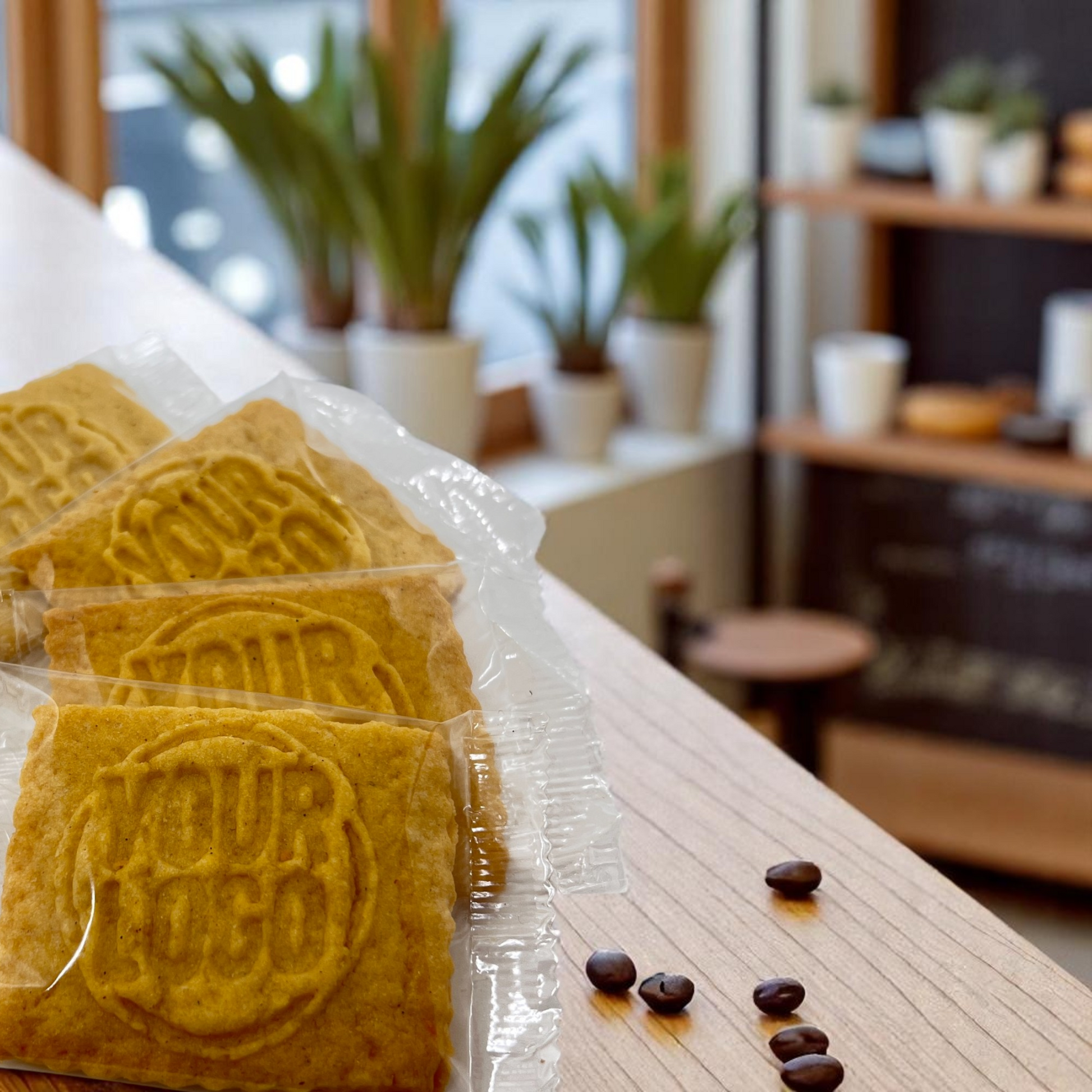 Einzeln verpackte Logokekse mit individueller Logo-Prägung, arrangiert auf einer Thekenkante mit Kaffeebohnen – ideal als Kaffeebeilage oder Kunden-Giveaway im Café