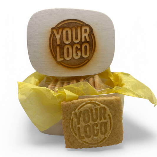 Holzkästchen mit gelasertem Logo im Deckel, gefüllt mit 8 Logokeksen in gelbem Seidenpapier – ein Keks zur Ansicht vorn platziert